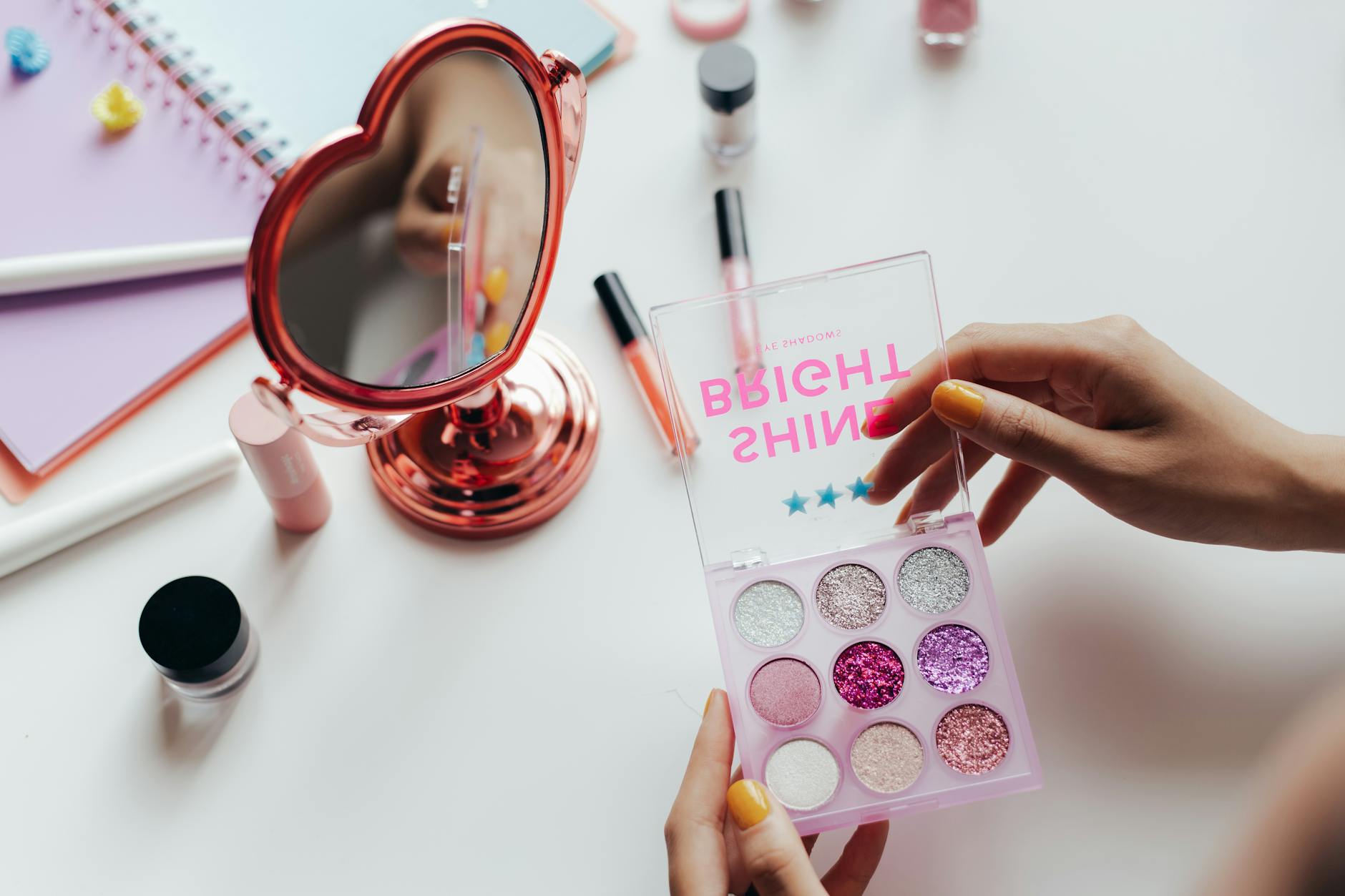 A vibrant eyeshadow palette with glittery colors held near a heart-shaped mirror.