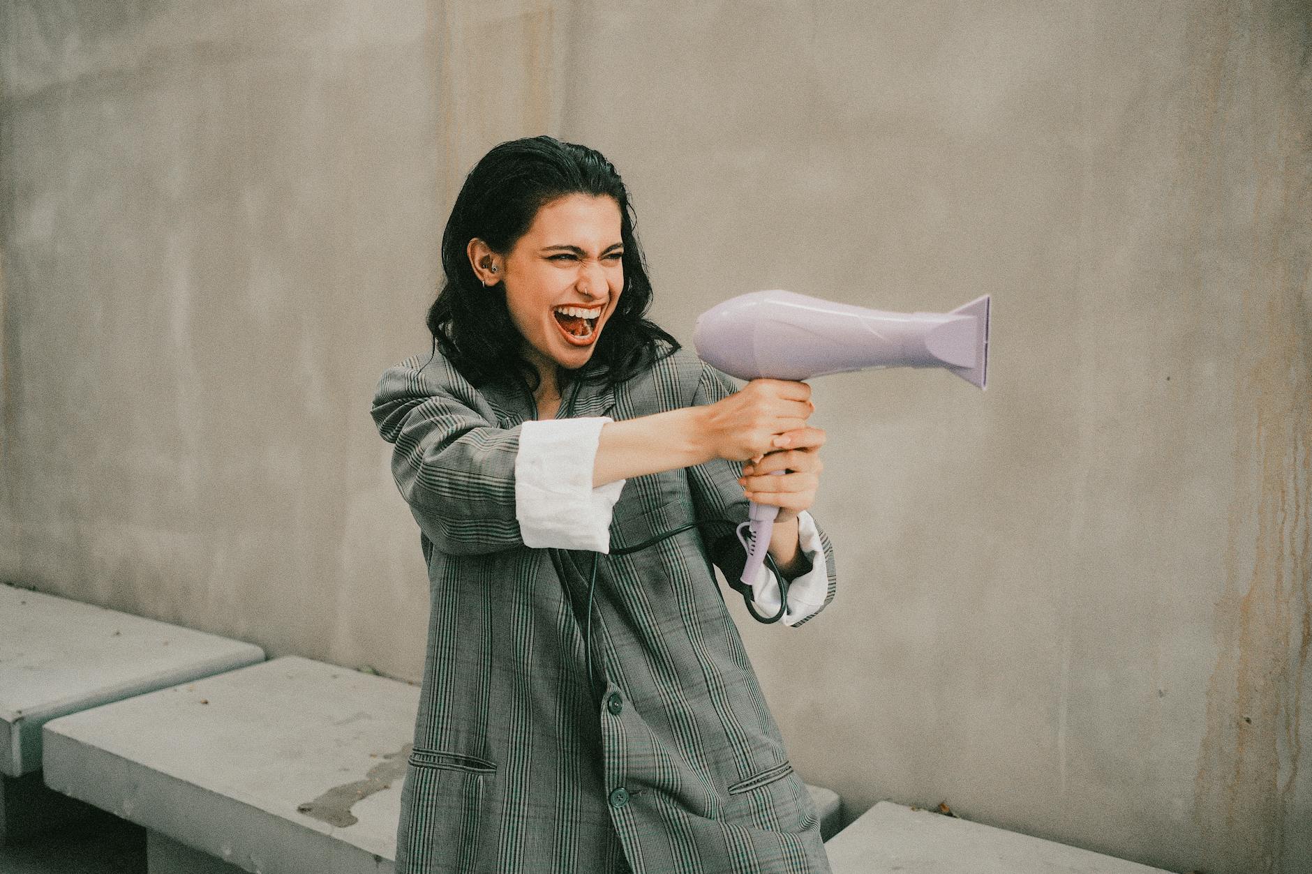 Joyful woman in oversized coat holding a hair dryer, mimicking a playful pose.