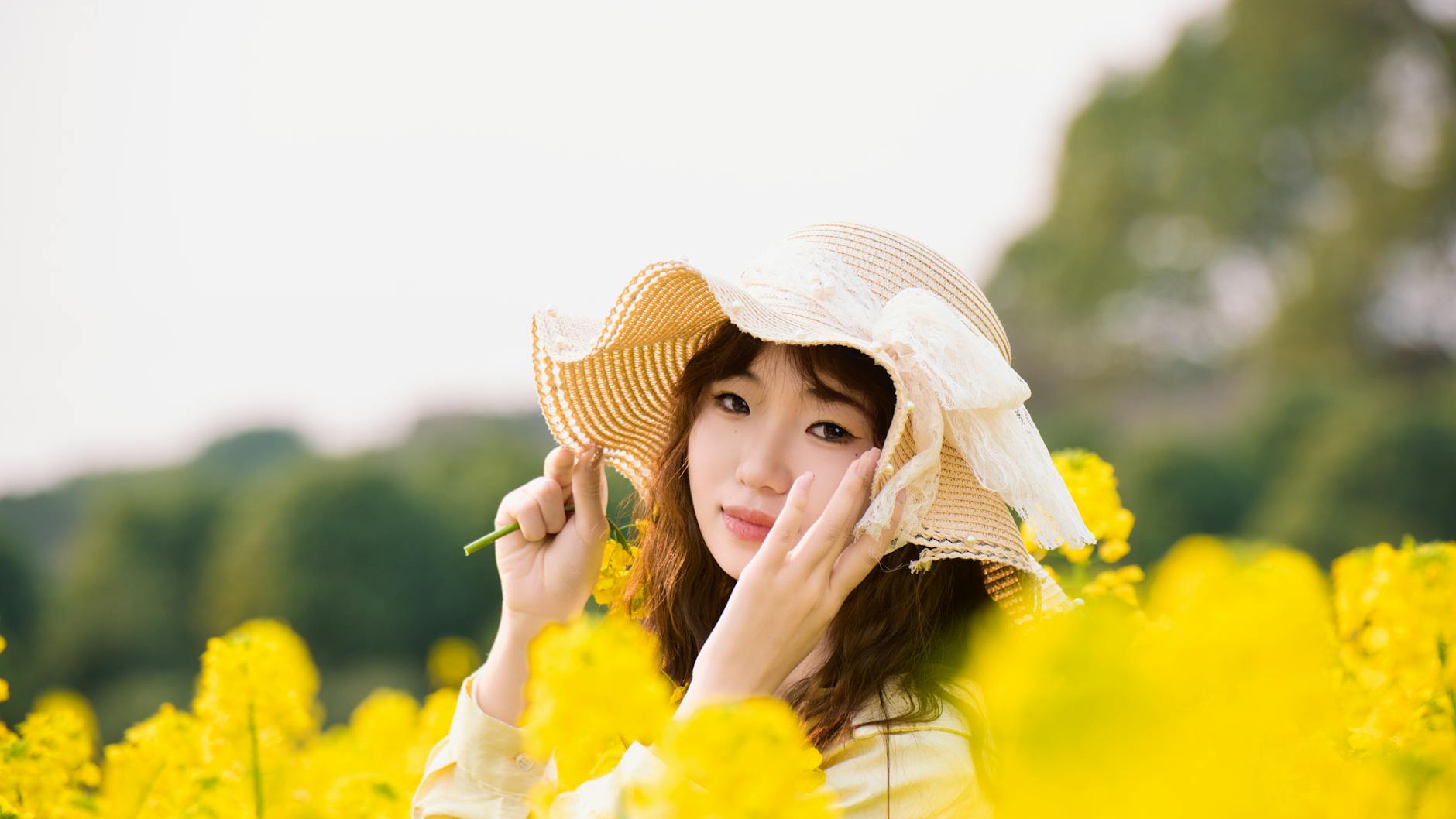 Young woman wearing a straw hat surrounded by bright yellow flowers outdoors.