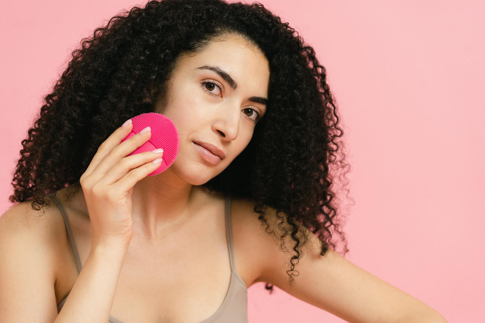 Portrait of a woman using a facial cleansing brush against a pink background.